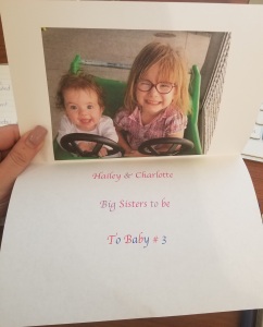 Photo of a card. The top is a picture of Hailey and Charlotte. The bottom reads "Hailey and Charlotte. Big sisters to be. To baby number three.".