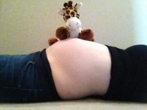 Photo of Melissa laying on her back showing the profile view of her pregnant belly. There is a gifaffe stuffed animal resting on her stomach.